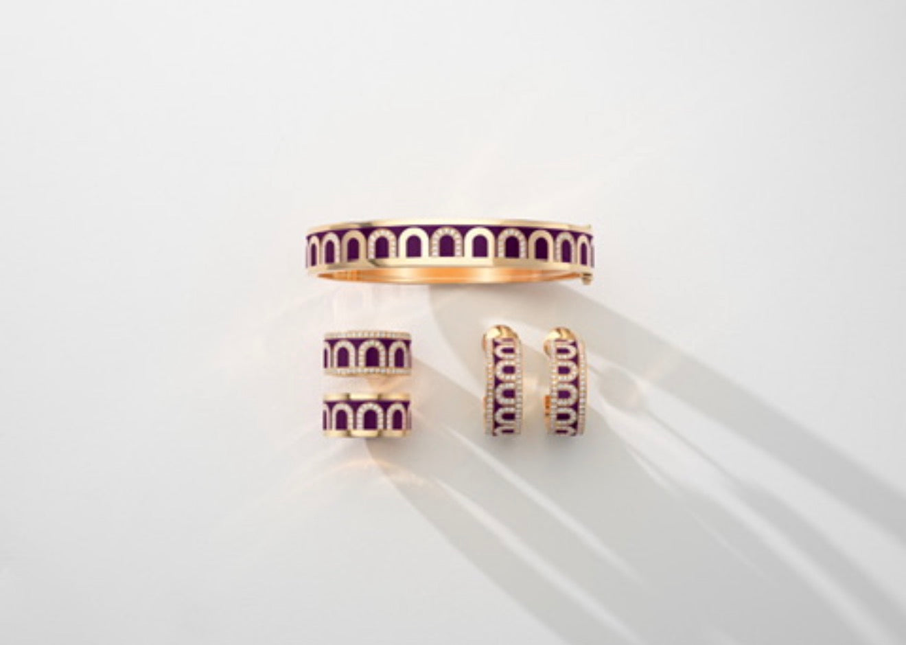 Gold jewelry set against a white background, featuring a bangle and matching hoops and rings with an arched purple and gold design, casting soft shadows to the right.