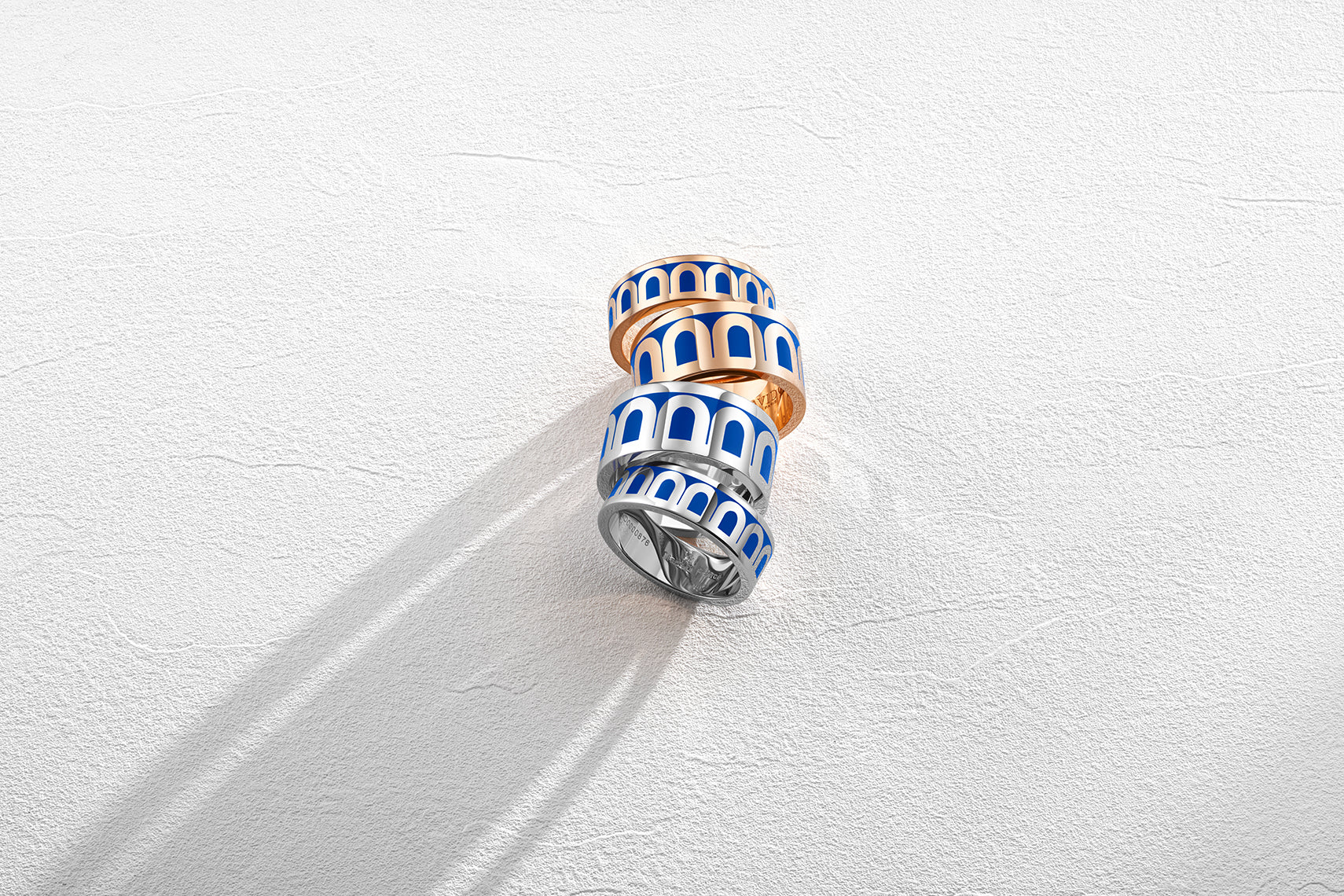 Three stacked rings with alternating gold and silver bands and a blue and white patterned design are placed on a textured white surface, casting subtle shadows.