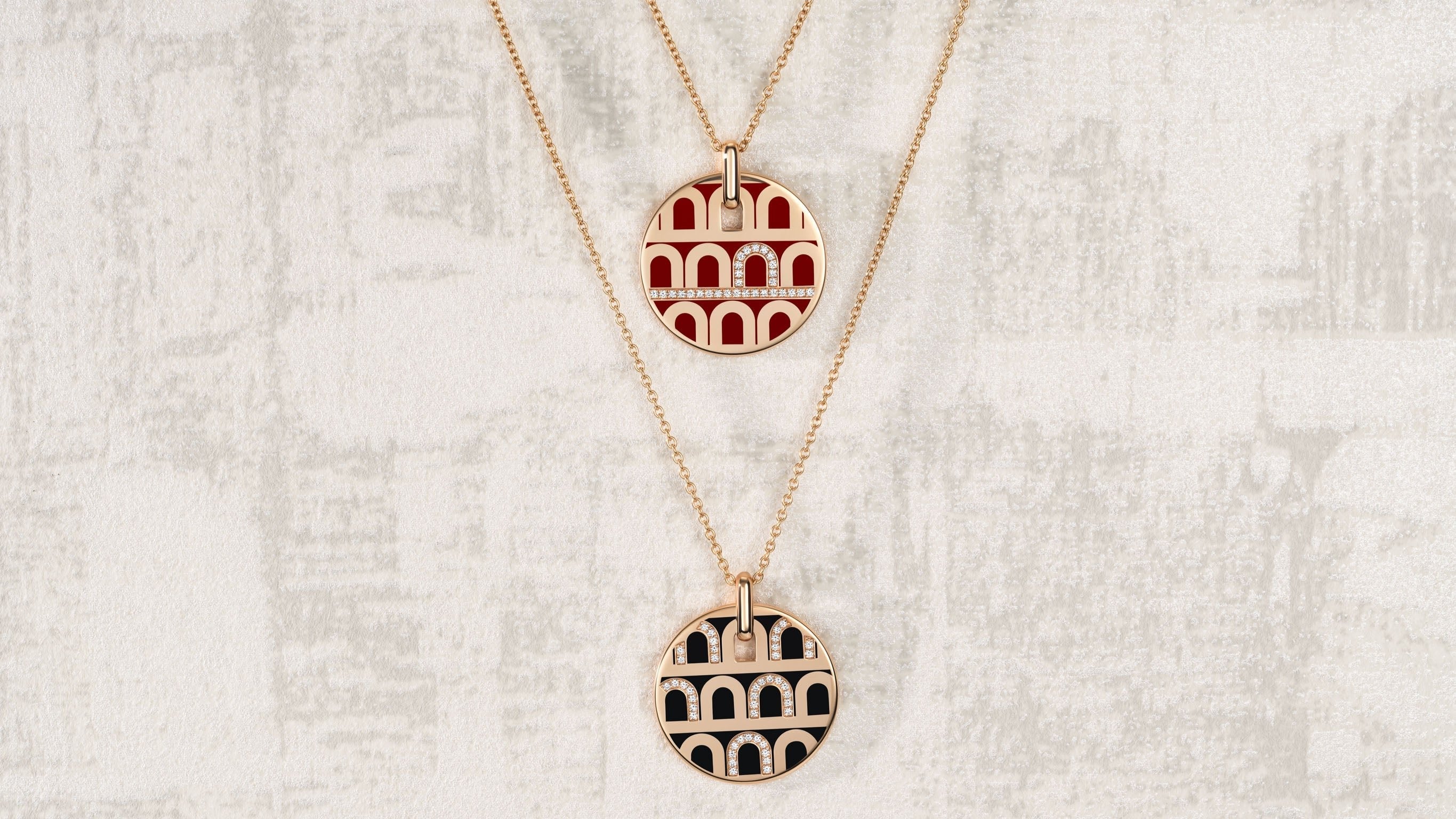 Two gold necklaces with round pendants featuring Colosseum designs, one in red and the other in black, are displayed against a textured, light-colored background.