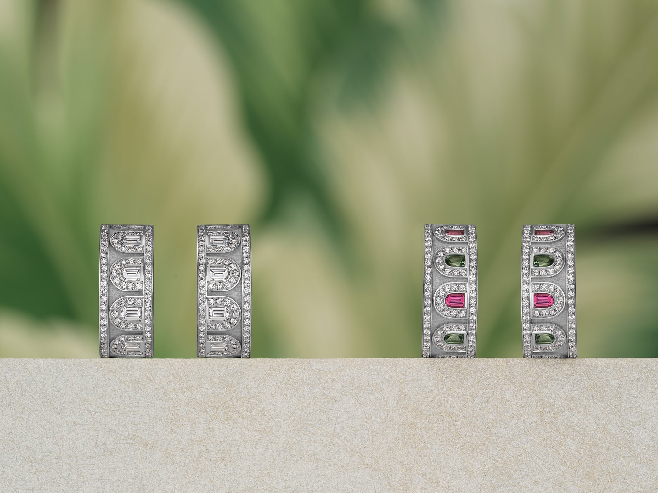 Four elegant rings with silver bands and intricate designs are displayed against a blurred green background. Some rings feature colorful gemstones in pink and green.