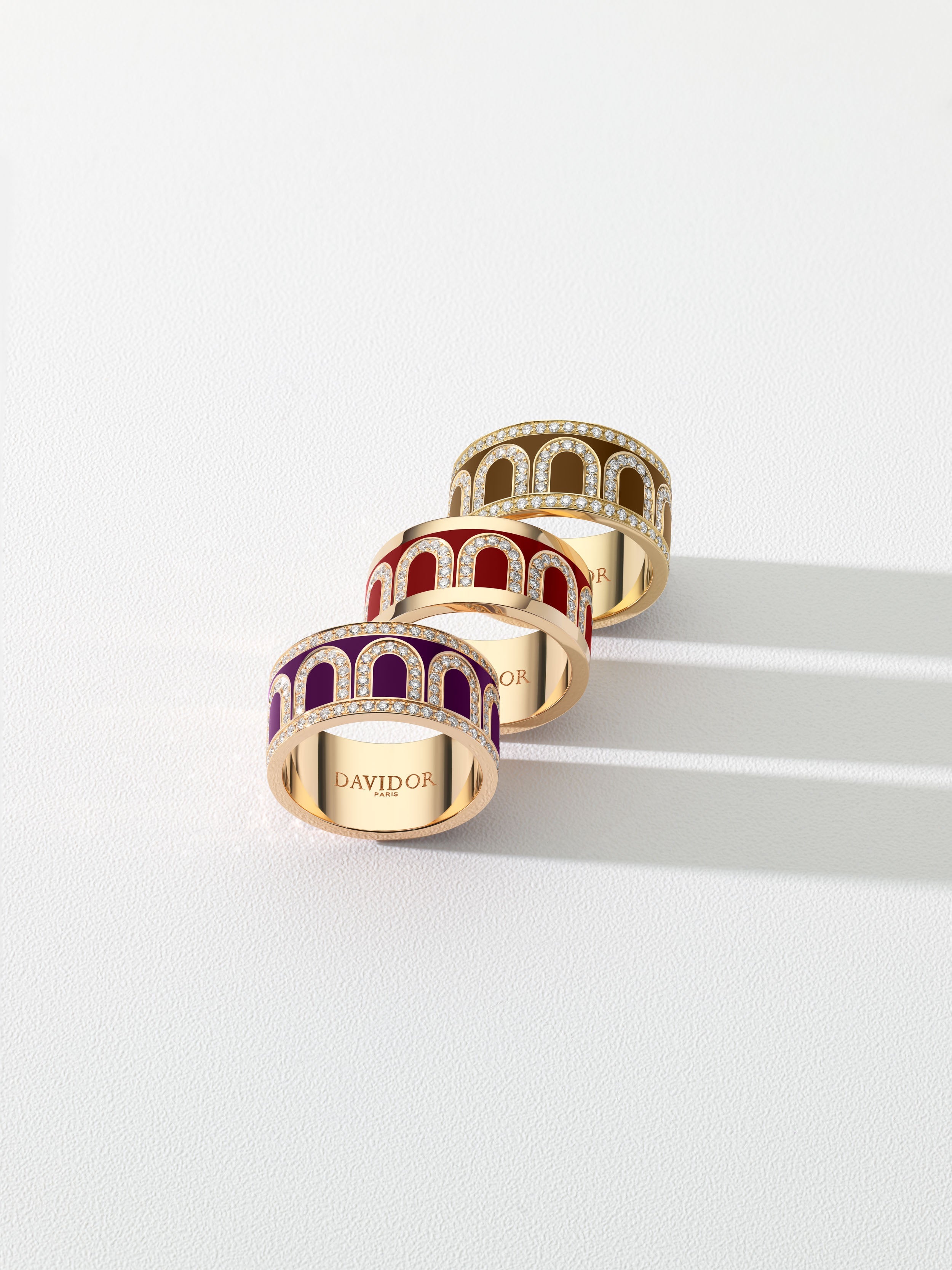 Six gold rings with intricate arch designs in brown, pink, and gray enamel, arranged in two rows on a light textured surface; “DAVIDOR” engraved inside each ring.
