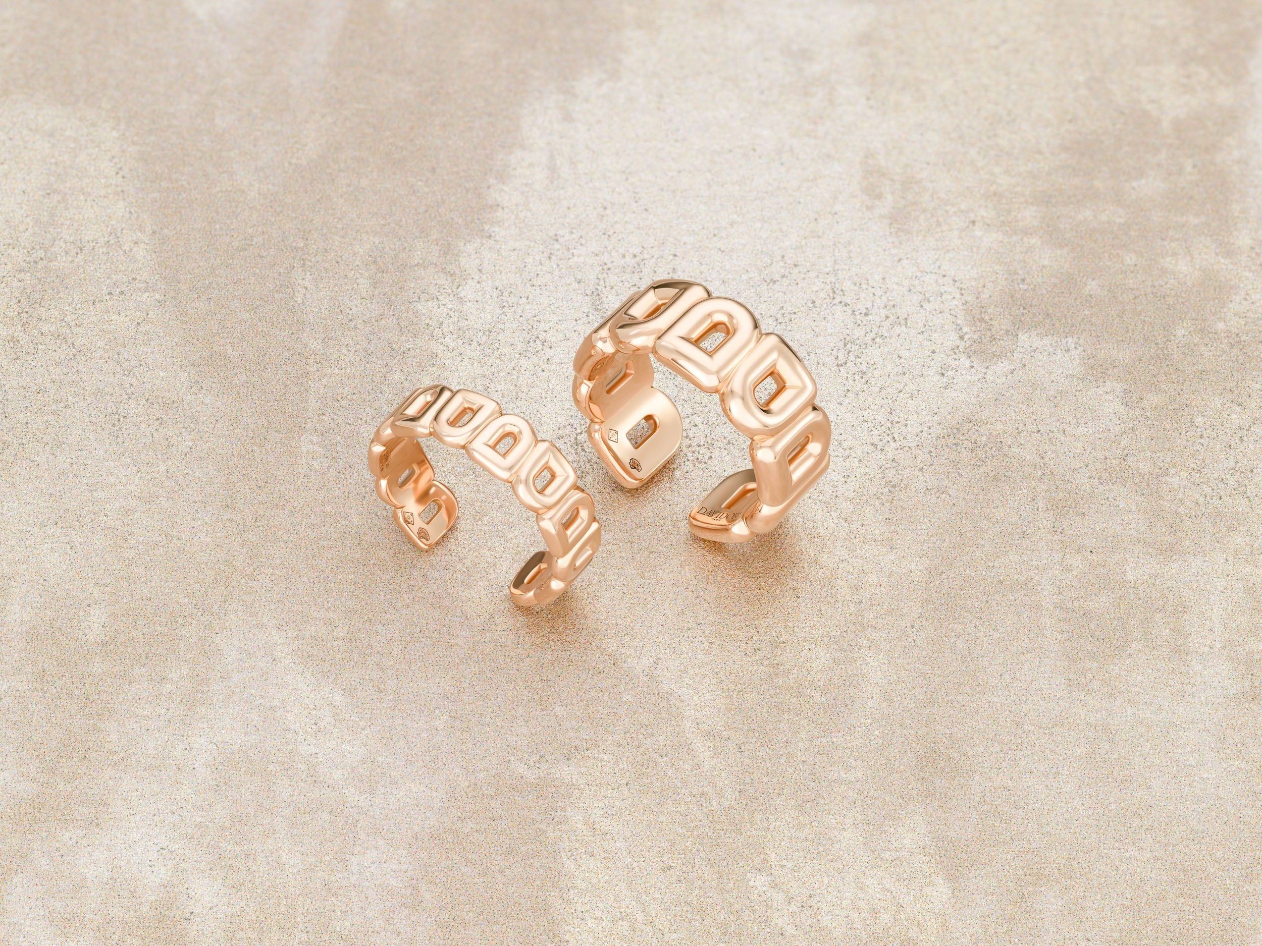 Two open-ended gold rings with geometric cutout designs are displayed on a light beige, textured surface. The rings are positioned side by side, with one slightly larger than the other.