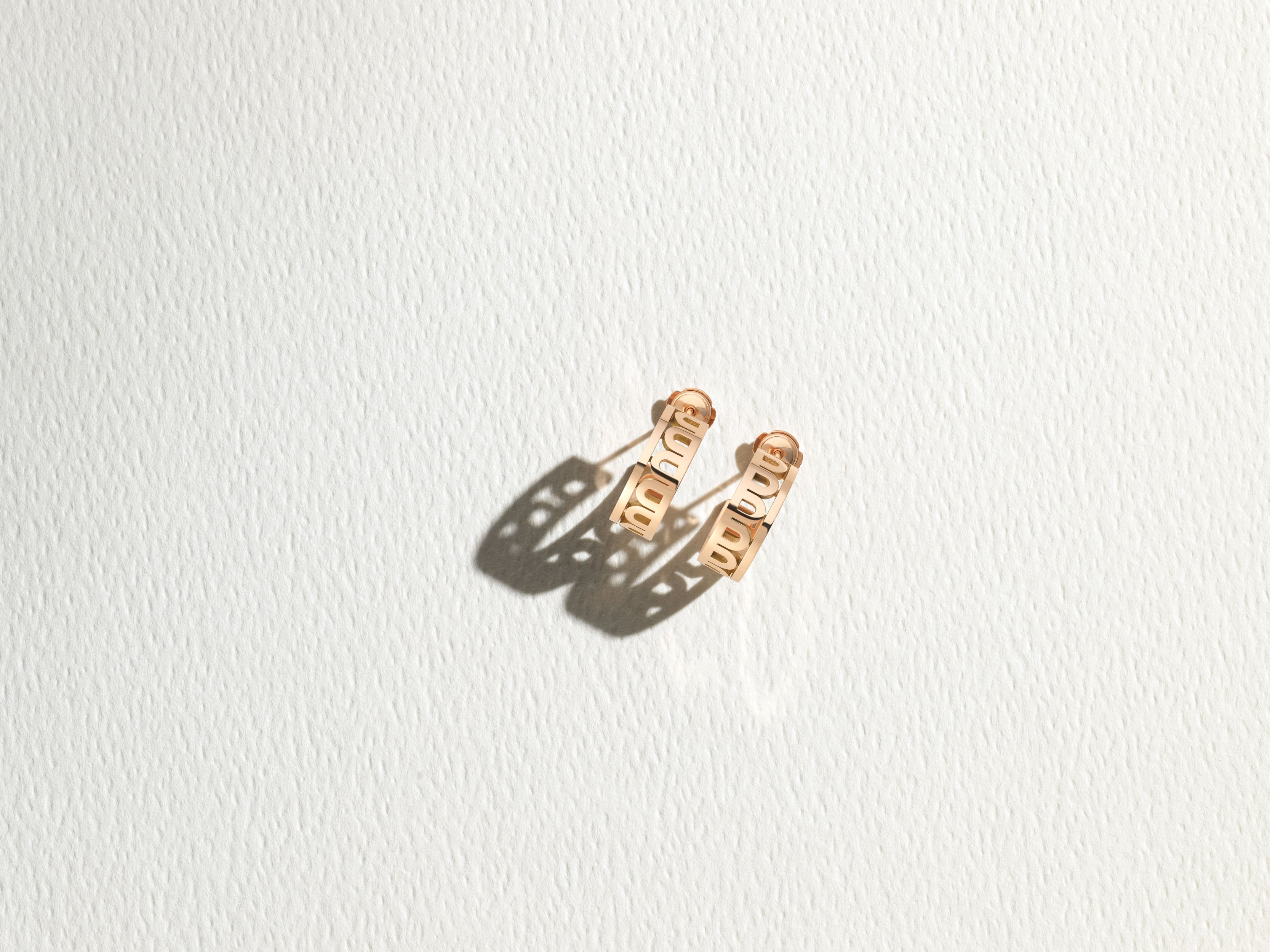 A pair of gold earrings with cut-out designs casting distinct shadows sits on a textured white surface, illuminated by direct light.