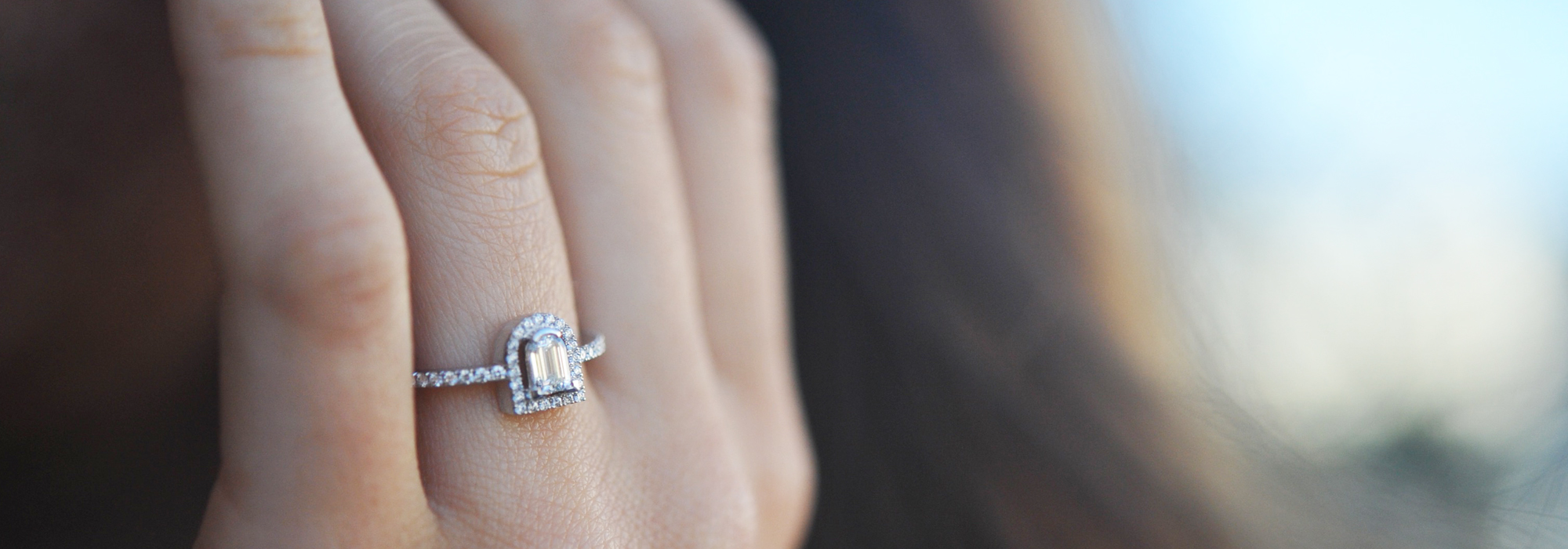 Close-up of a persons hand wearing a diamond ring with an elegant design. The background is softly blurred, highlighting the details of the ring.