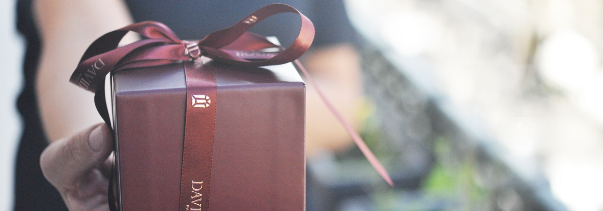 A person holds out a wrapped gift box adorned with a dark red ribbon. The box has elegant branding, and the background is softly blurred, emphasizing the gift.