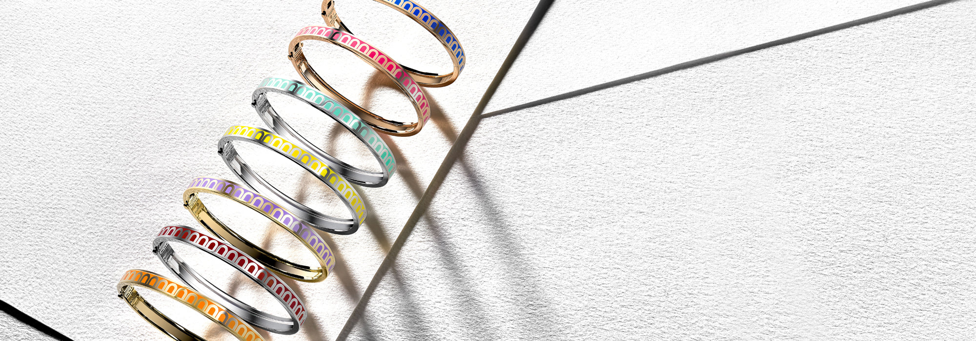 A collection of seven elegant jewelry bangles in various pastel colors, including pink, orange, blue, green, yellow, purple, and gray, arranged diagonally on a textured white surface. Shadows fall across the background.