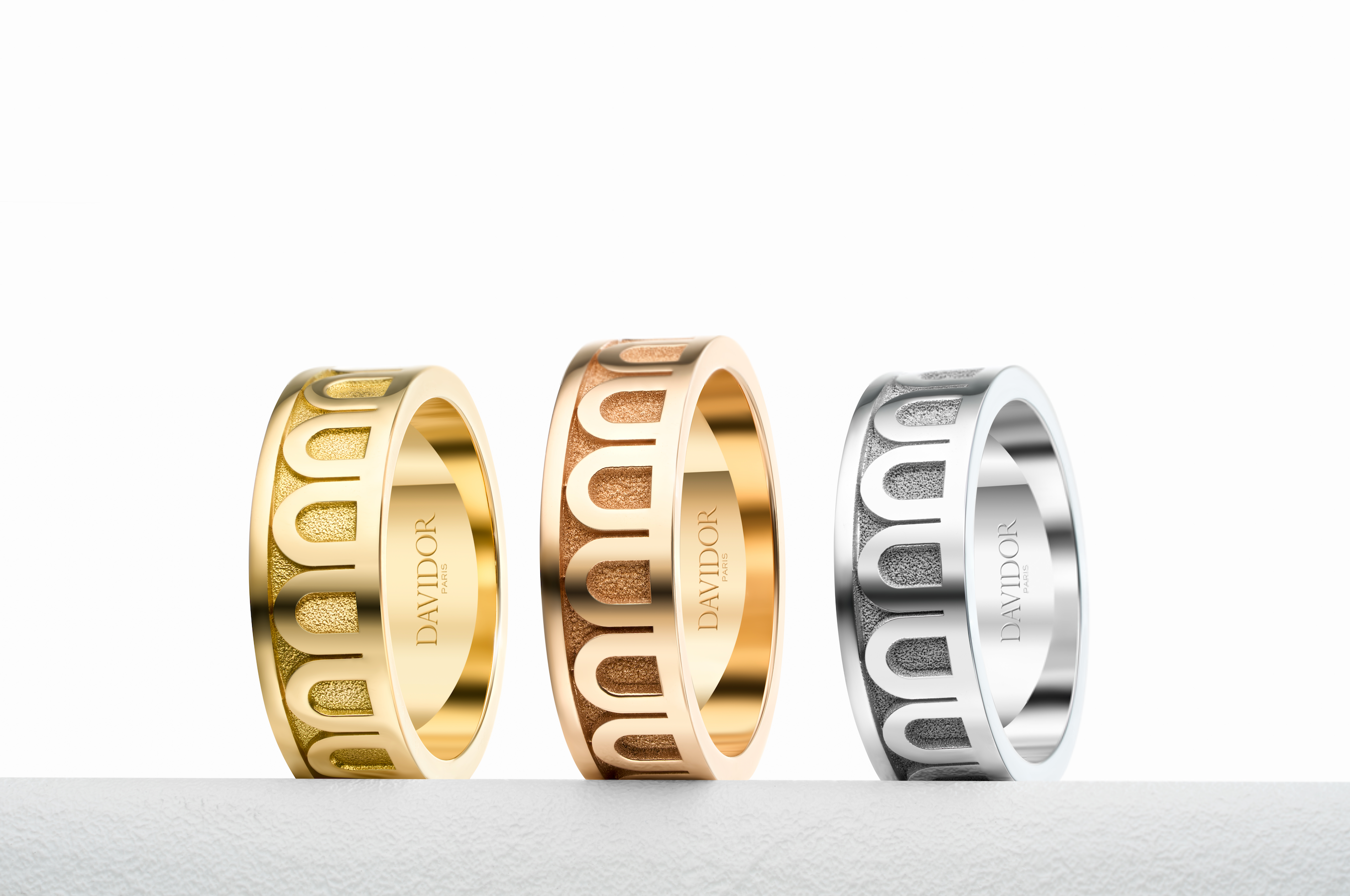 Three DAVIDOR rings are displayed upright in a row: one gold, one rose gold, and one silver, each featuring an arch pattern and engraved branding on a white background.