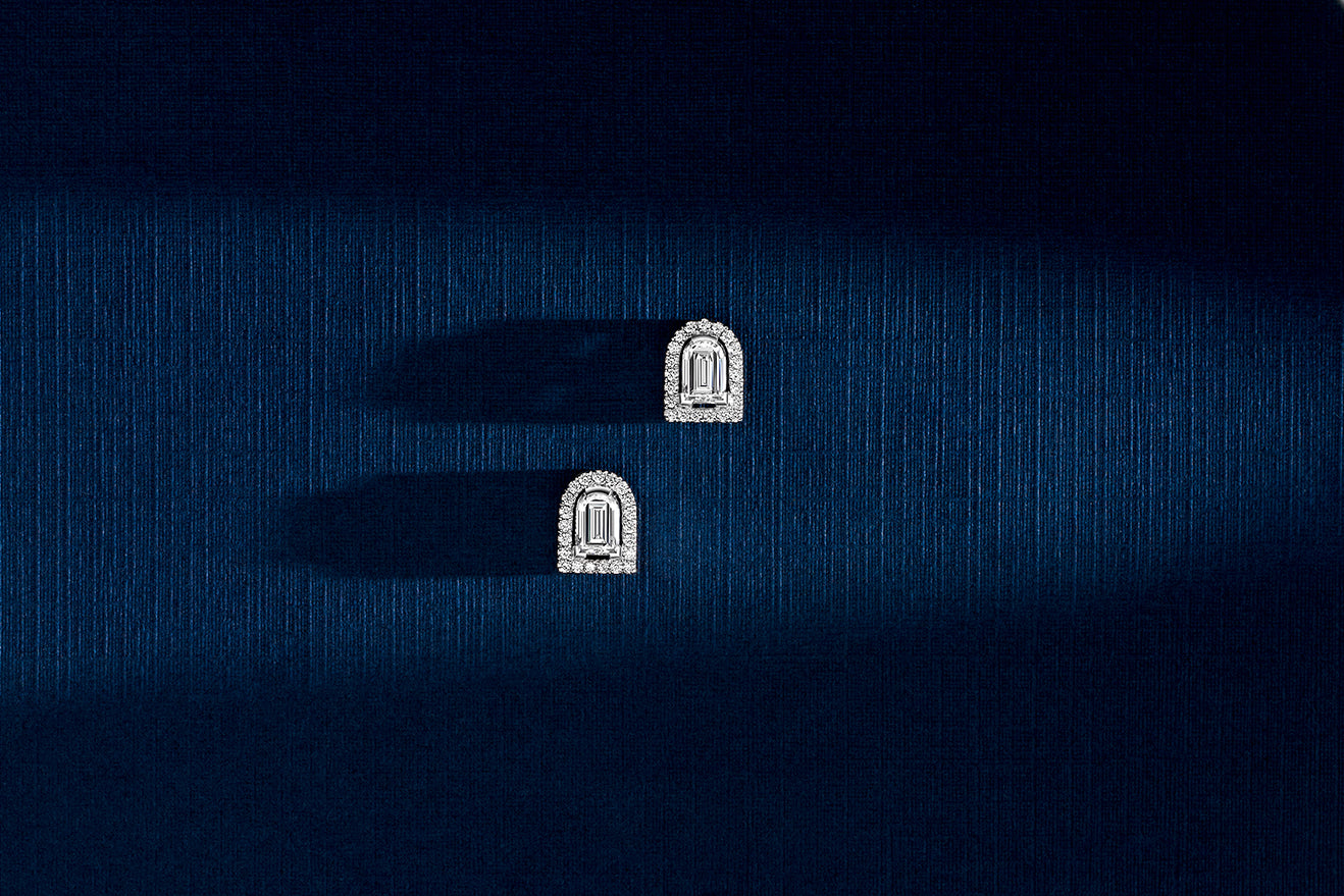 Two ornate, rectangular diamond earrings with rounded tops are placed on a textured dark blue surface, casting long shadows to the left.