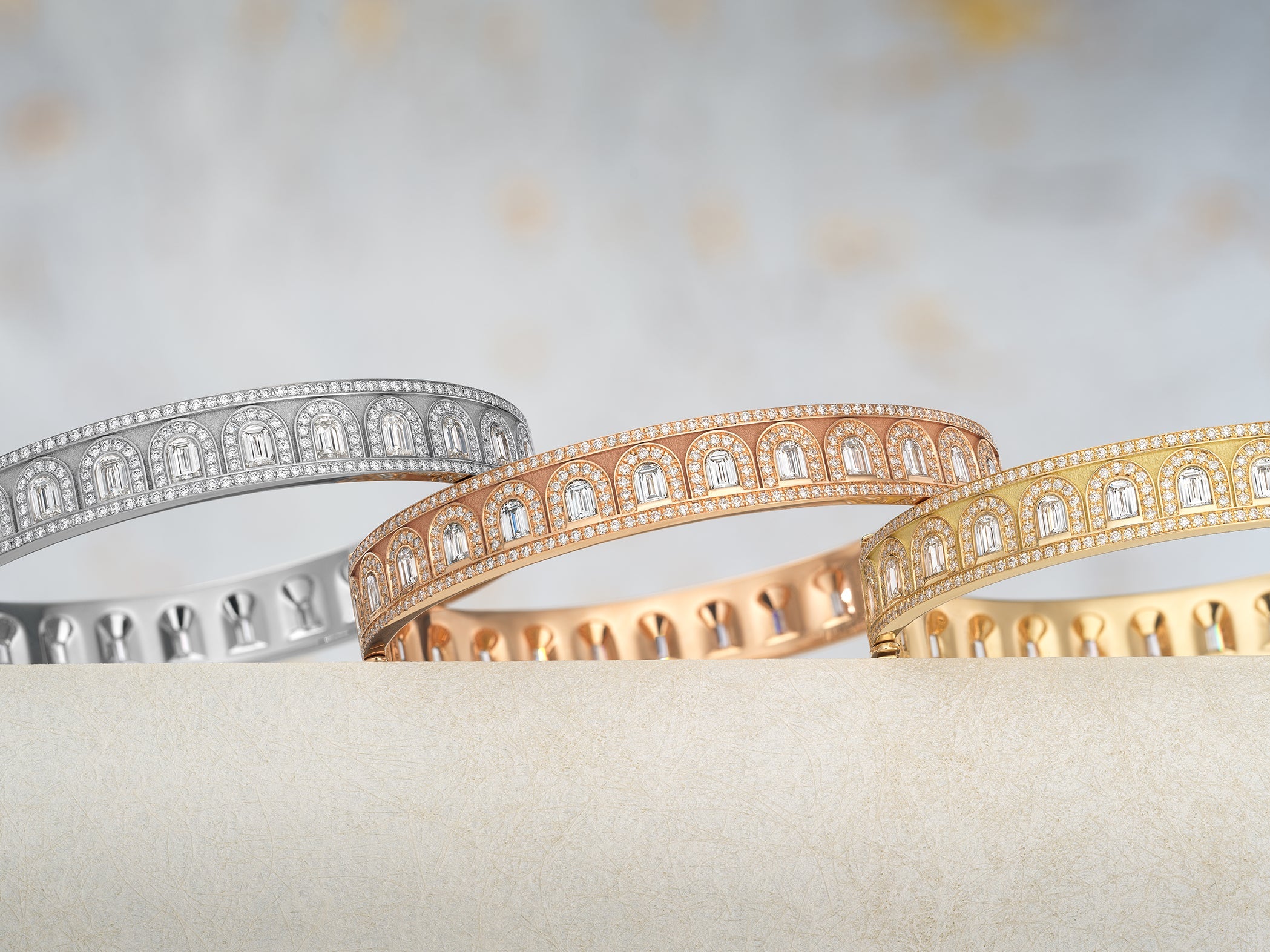 Three circular rings with intricate designs are placed on a textured, light-colored surface. The rings are gold, silver, and rose gold, each featuring small, square patterns around the band.