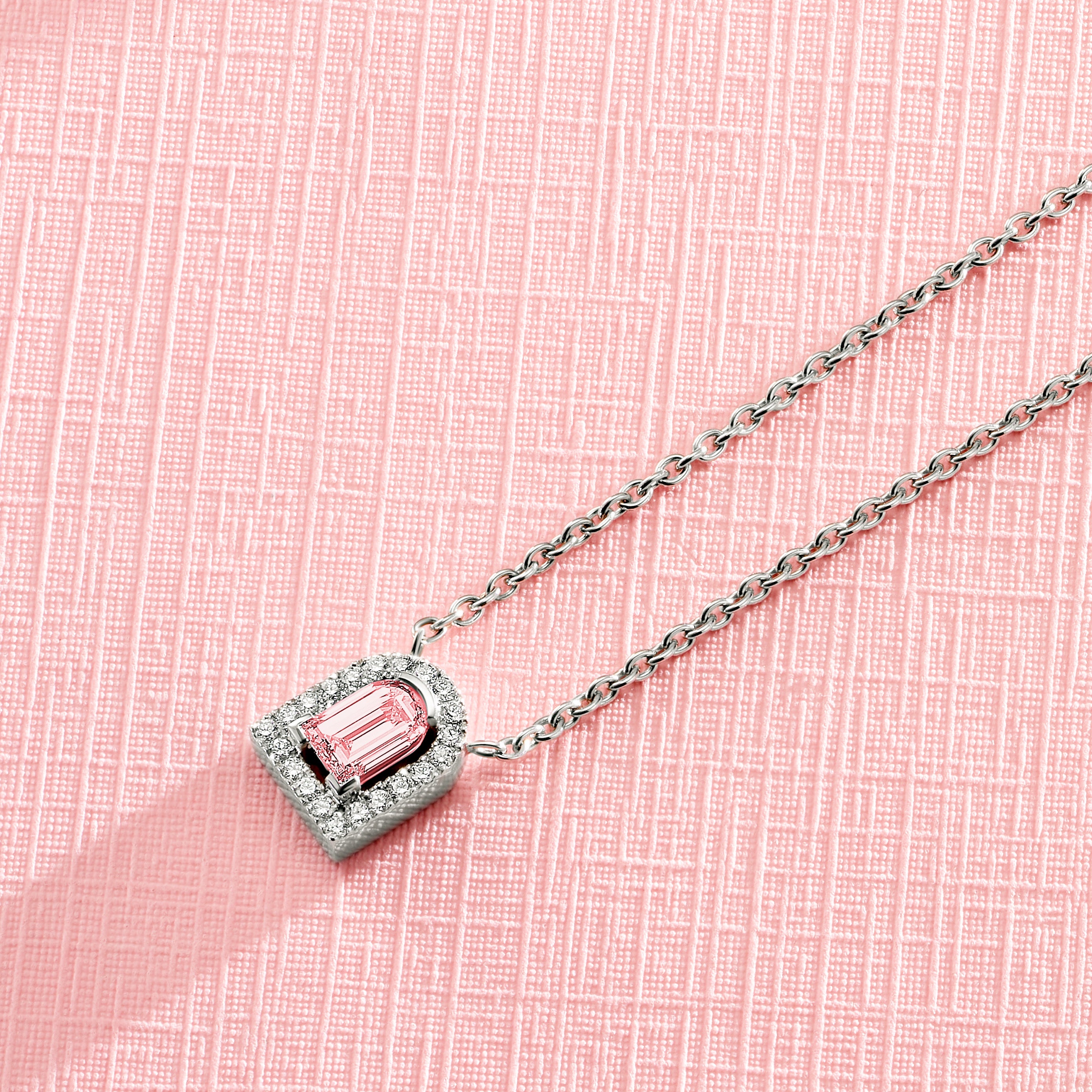 A silver necklace with a rectangular pink gemstone pendant surrounded by smaller crystals, placed on a pink textured background.