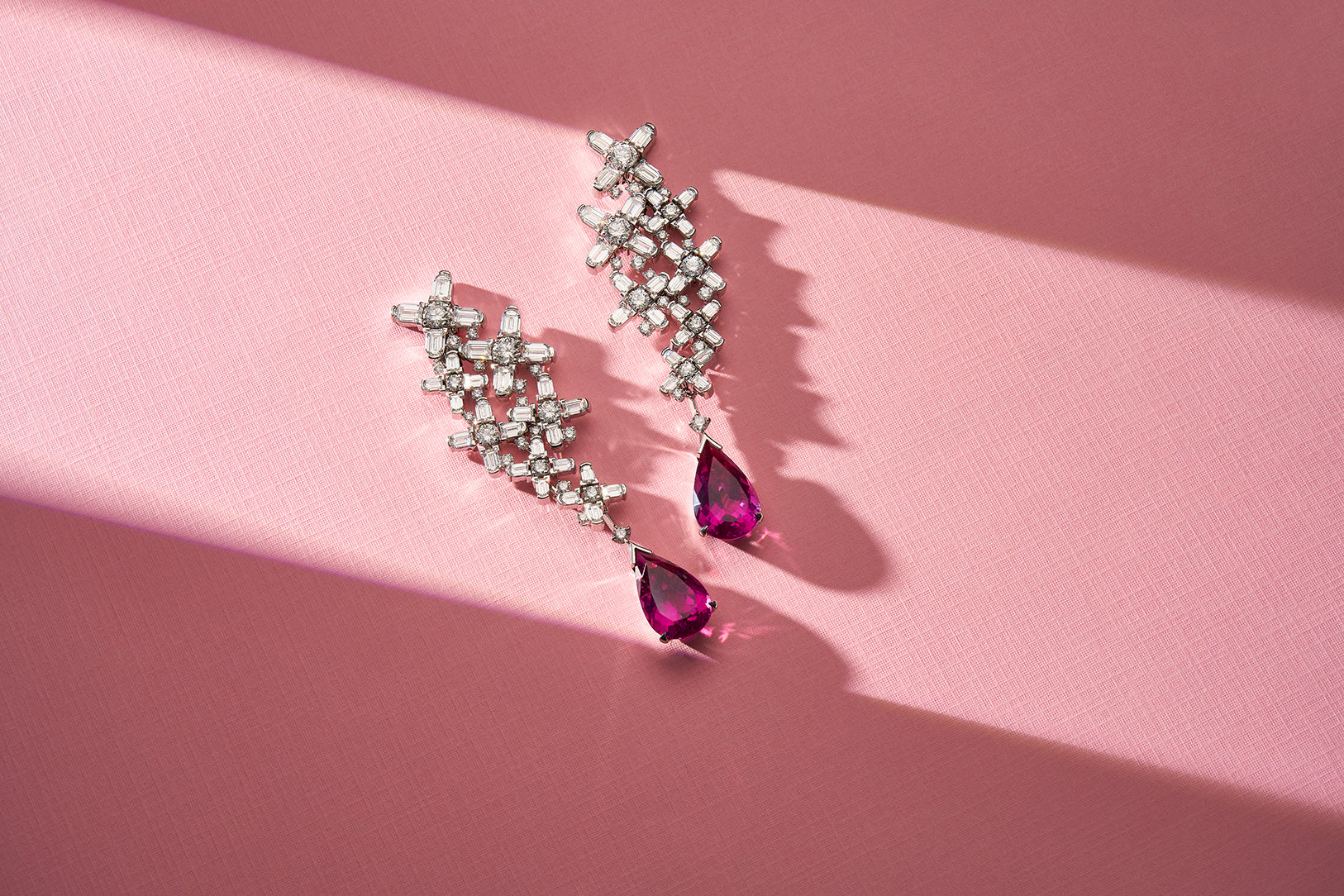 Elegant silver earrings with intricate geometric designs featuring clusters of small diamonds and teardrop-shaped pink gemstones at the ends, placed on a textured white surface.