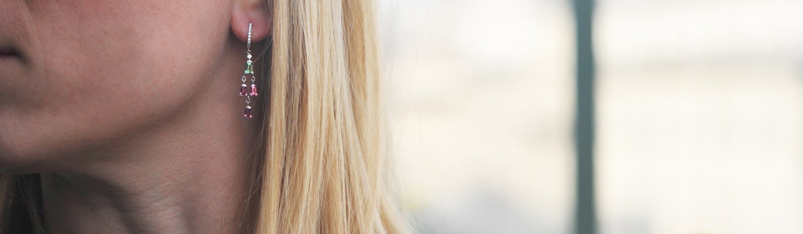 Close-up of a person with long blonde hair wearing dangling earrings that feature green and pink gemstones. The background is blurred.