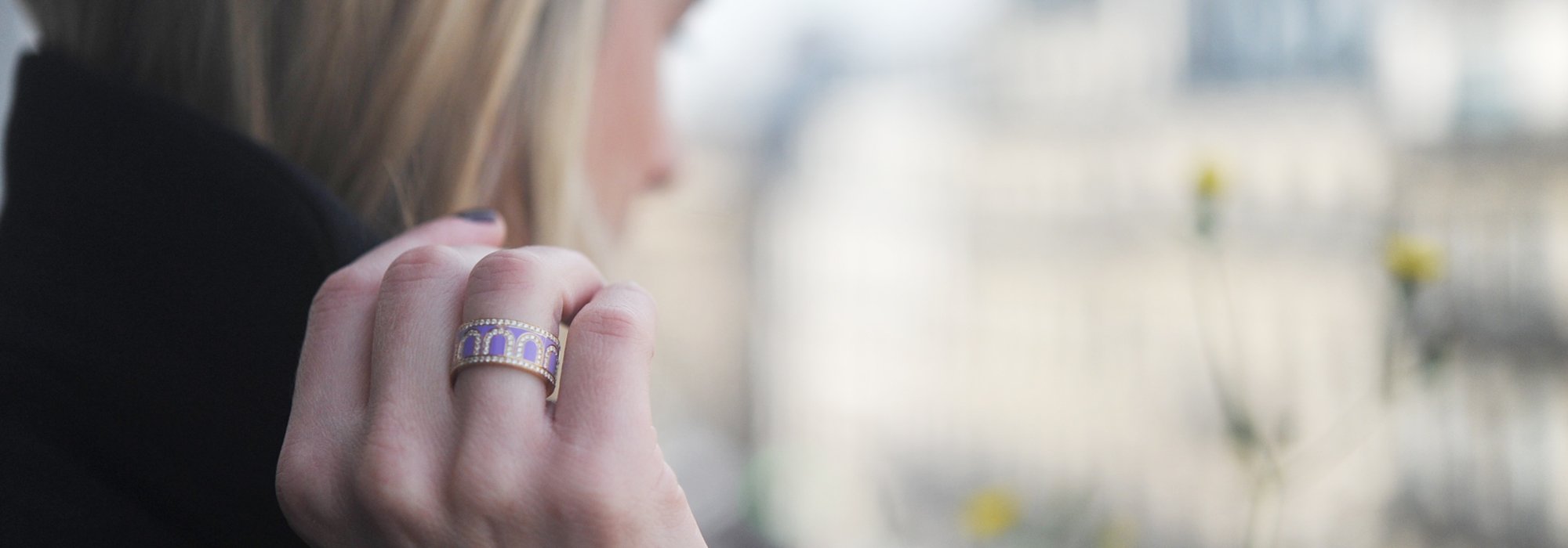 A person with blonde hair, seen from the side, is holding their chin. The focus is on a gold and purple patterned ring on their finger. The background is softly blurred cityscape.