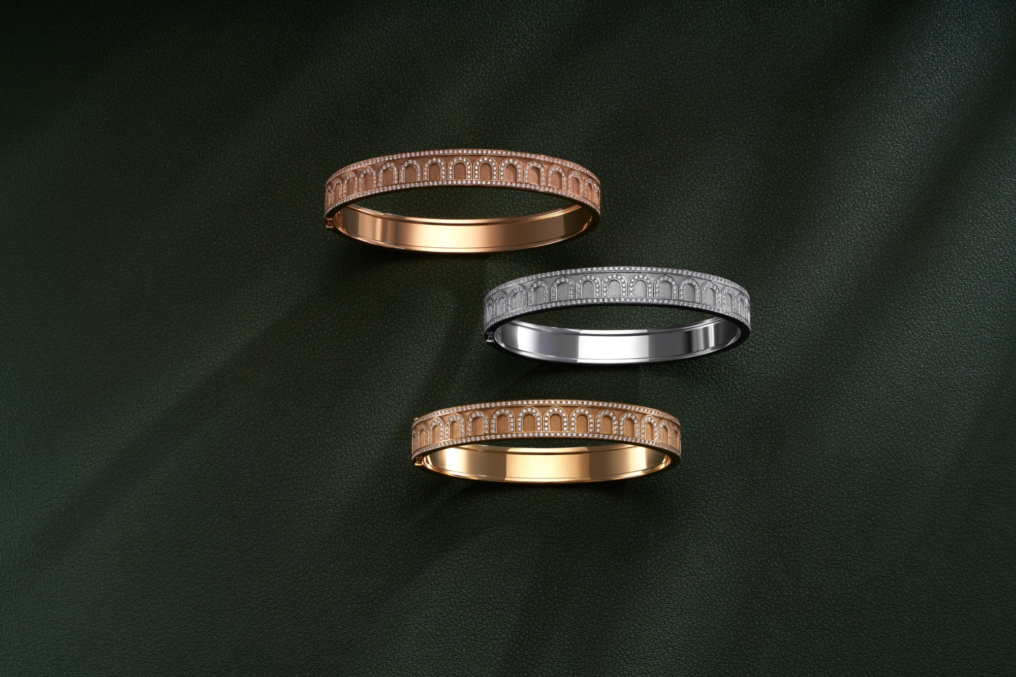 Three intricately designed rings are displayed on a dark green textured surface. The rings, in rose gold, yellow gold, and silver, feature detailed arch patterns, casting subtle shadows.