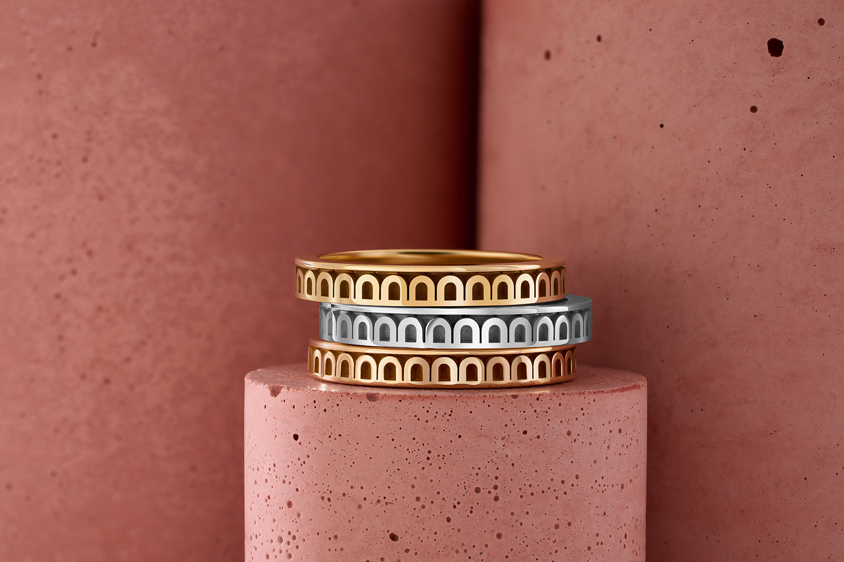 Three rings stacked on a textured pink surface, featuring gold and silver colors with arched designs. The background consists of similar pink textured surfaces.