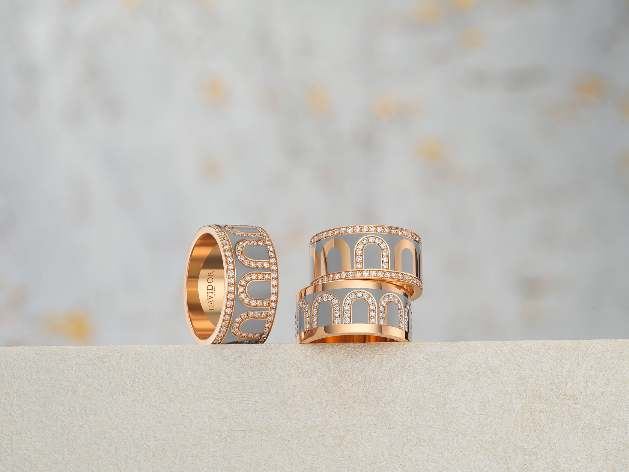 Two elegantly designed gold rings adorned with intricate patterns and small diamonds sit on a textured, light-colored surface against a blurred background, featuring subtle hints of gold and gray.
