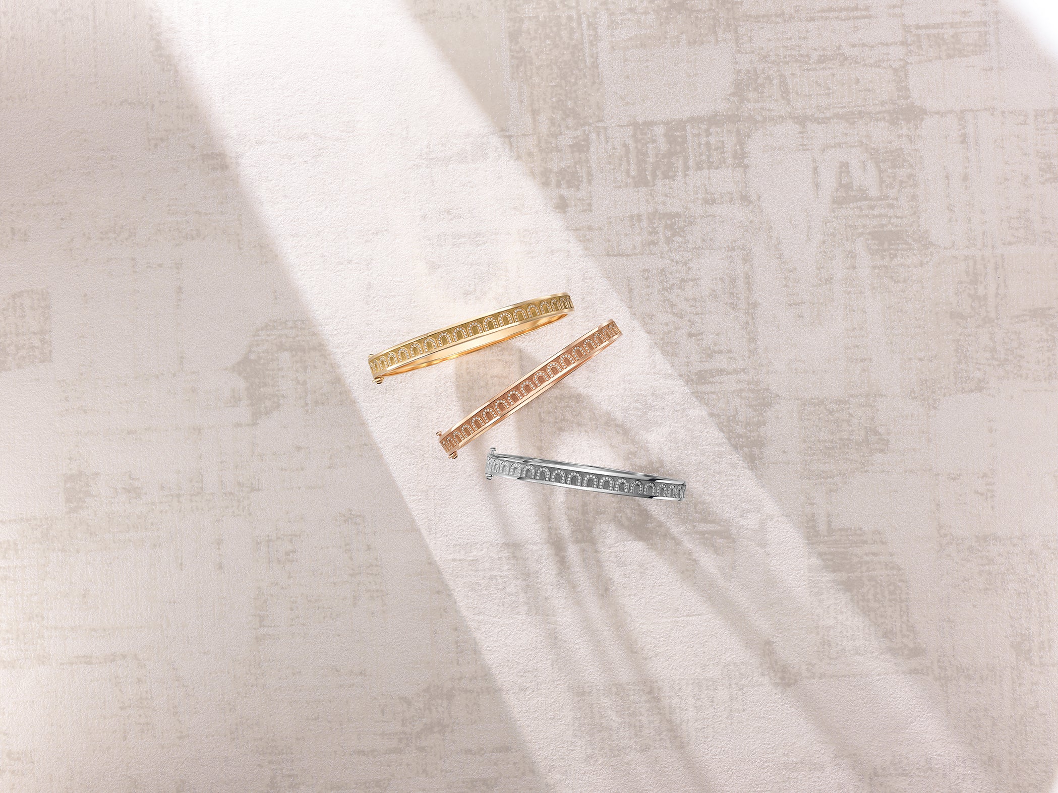 Three metal hair clips—gold, rose gold, and silver—are arranged diagonally on a textured white surface. Sunlight casts soft shadows across the scene, highlighting the clips intricate designs.