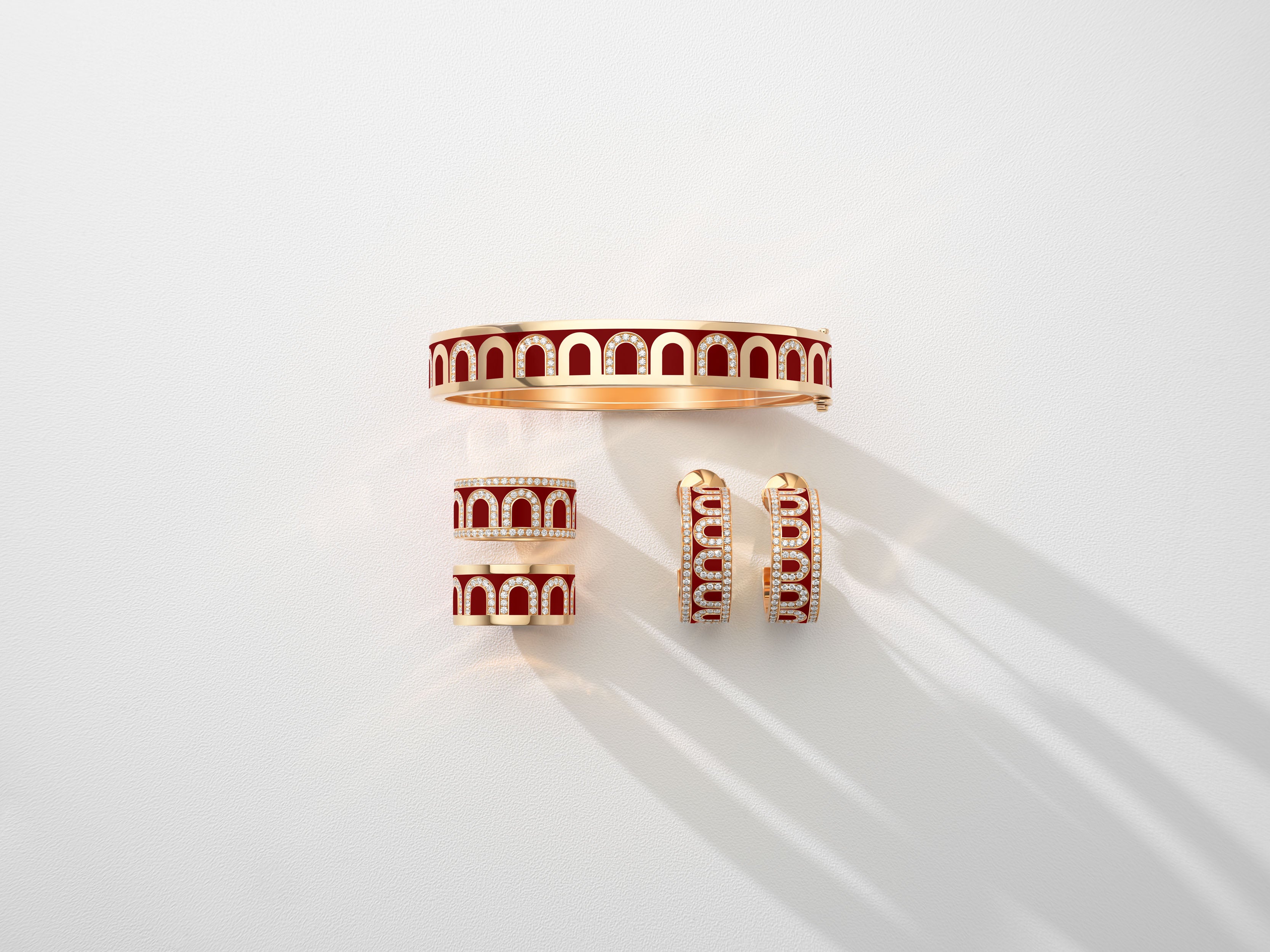 A L'Arc de DAVIDOR Bangle MM in 18k rose gold with lacquered ceramic and Colonnato diamonds, along with a wide ring and hoop earrings featuring red and white arch patterns, are arranged on a white surface casting soft shadows to the right.