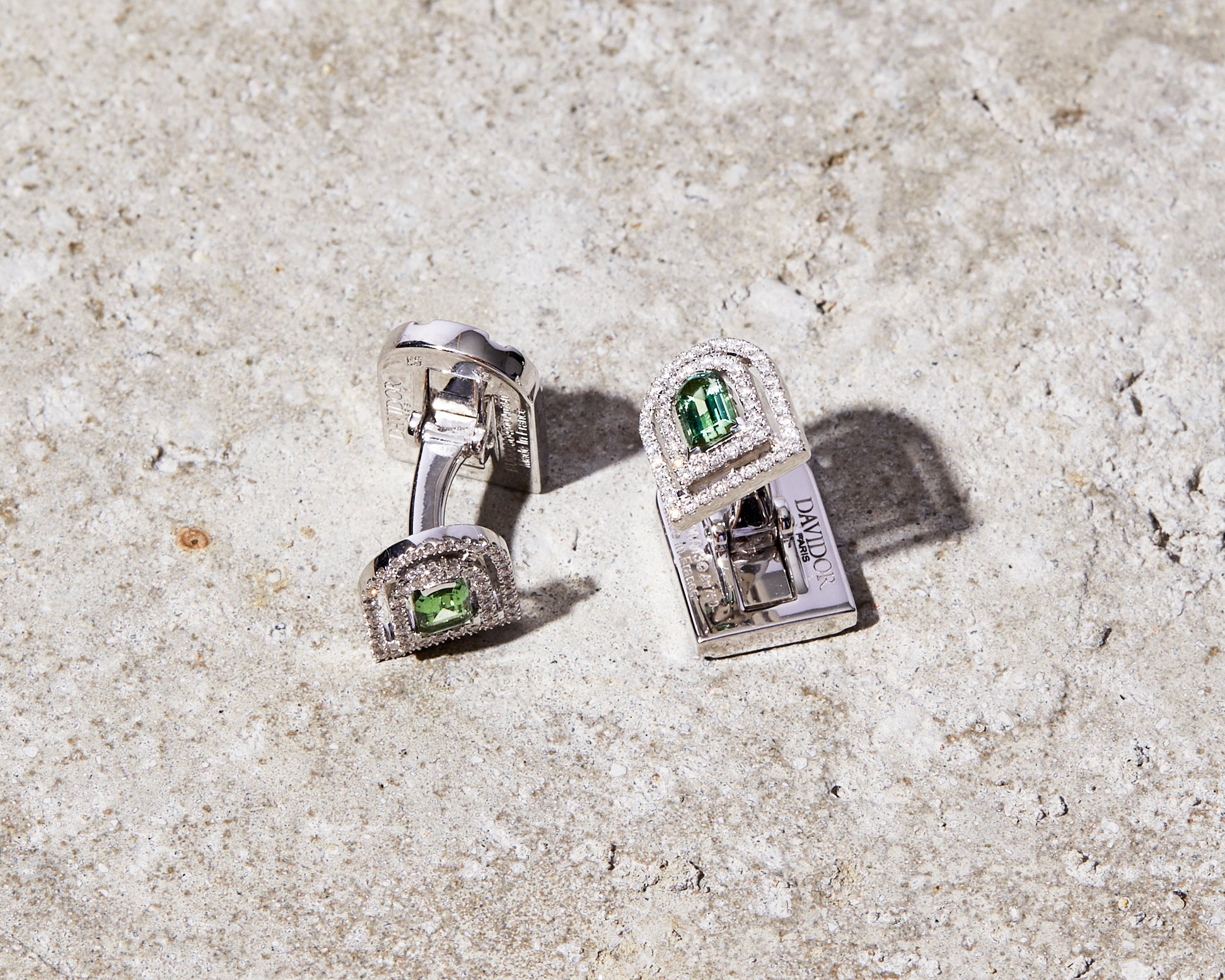 Two silver cufflinks with green gemstones and small white stones embedded, placed on a textured grey concrete surface. The cufflinks have a rectangular shape with a slightly raised design.