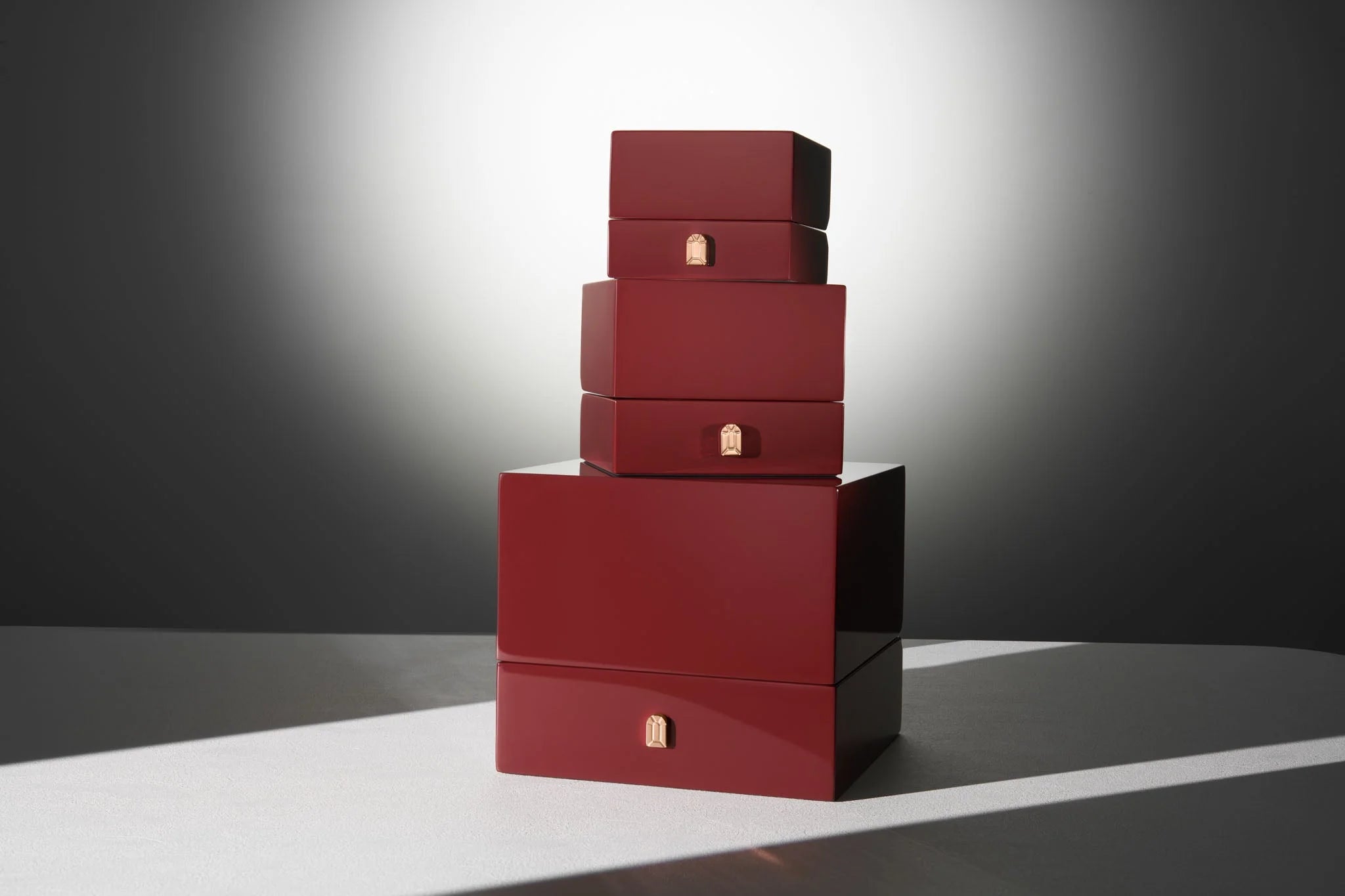 A stack of four dark red boxes of varying sizes on a smooth surface, with a spotlight effect in the background, highlighting their glossy finish. Each box has a small gold emblem on the front.