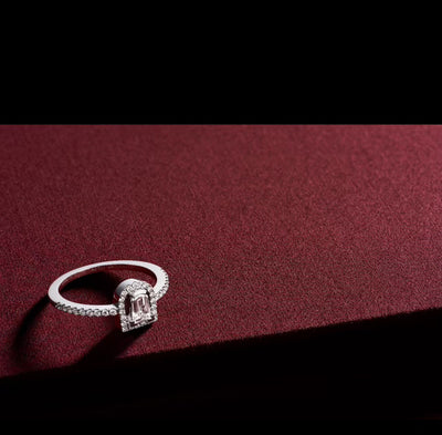 A diamond ring from DAVIDOR, featuring an emerald-cut center stone surrounded by a halo of smaller diamonds, rests on a textured deep burgundy surface. The contrasting black background enhances the elegance and sparkle of the TEST 1 ring.