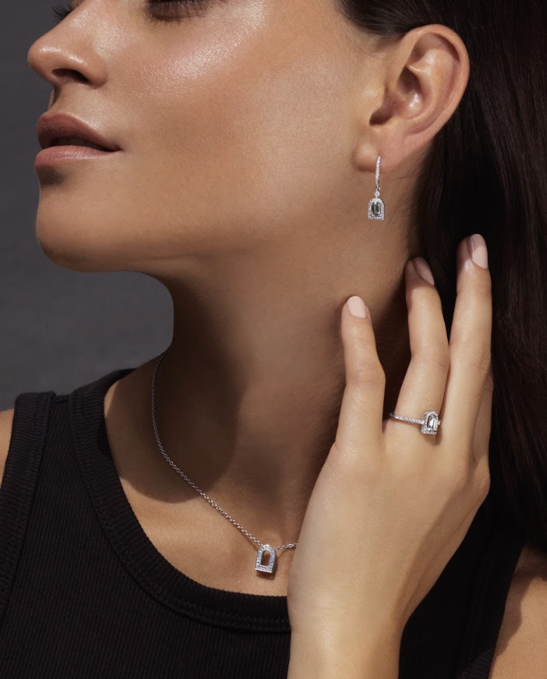 A woman wearing a black top showcases matching jewelry: a necklace, earrings, and a ring, each featuring a rectangular gemstone with a border of smaller stones. She poses with one hand near her face against a dark background.